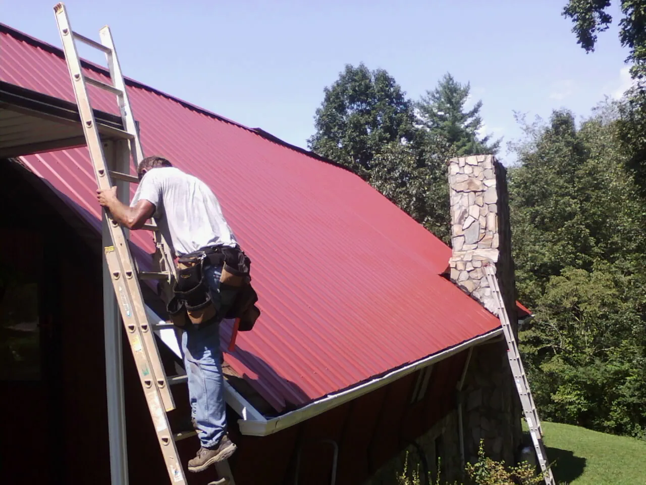Roofer on a ladder beside a red metal roof and stone chimney — Soffit & Fascia Repair by Roofing Brothers in Big Timber